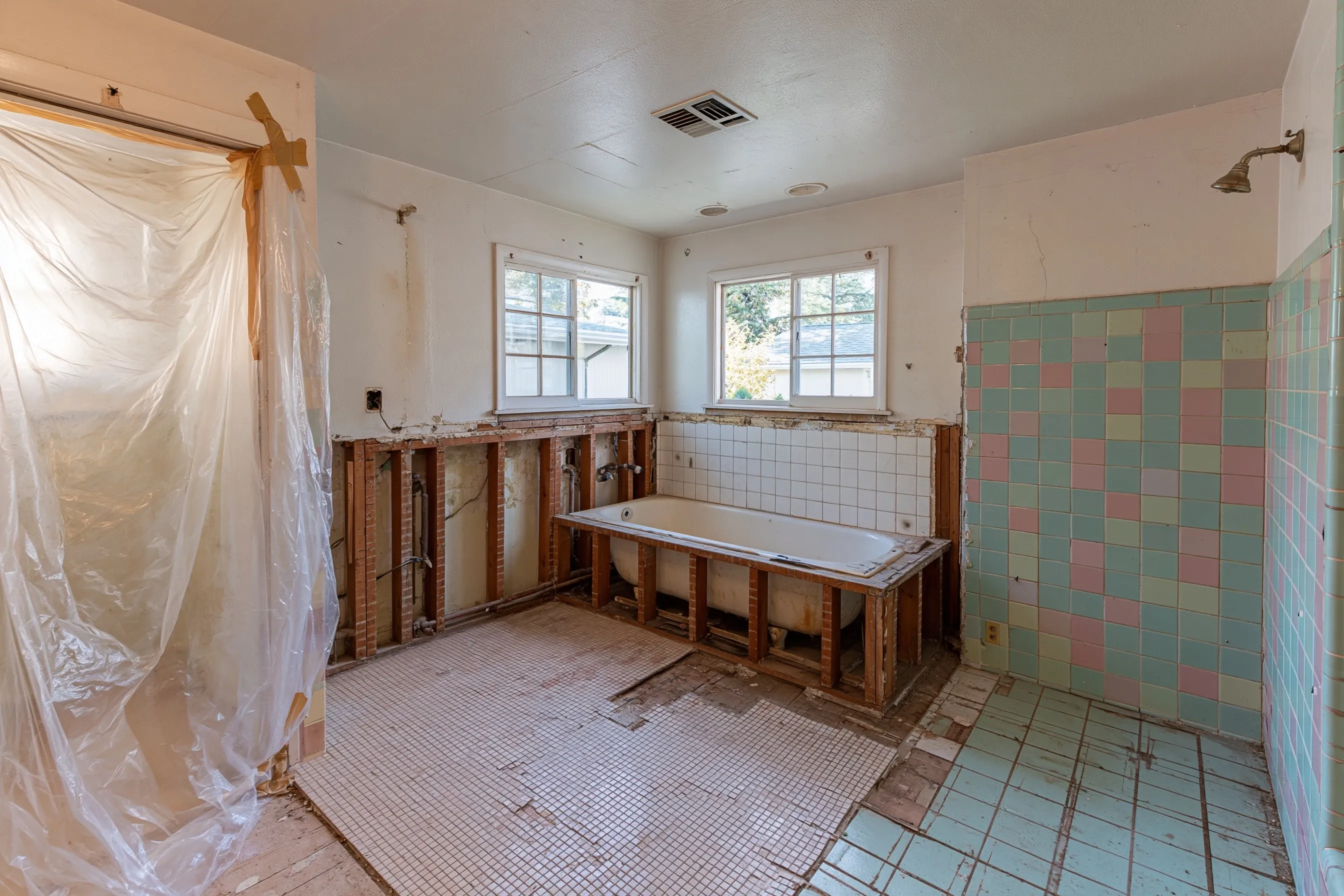 A 1950s bathroom opened for a remodel: tub-surround wall cut out, floor tile lifted, plastic up